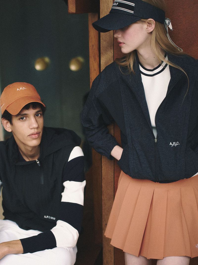 Two models wearing A.P.C. golf apparel including caps, a dark jacket, white polo, and an orange pleated skirt.