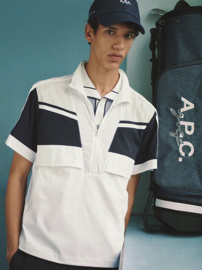 Model wearing a white and navy half-zip short-sleeve shirt and cap next to an A.P.C. golf bag against a blue backdrop.