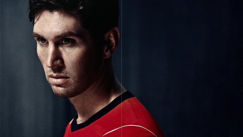 Dramatic close-up portrait of footballer John Stones wearing a red Nike England soccer jersey against a dark background.