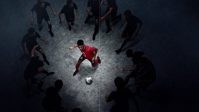 Soccer player in a red England kit dribbling a football inside a dramatic spotlight surrounded by shadowed players in black.