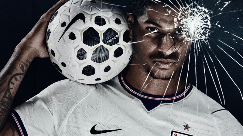 Marcus Rashford holding a Nike soccer ball on his shoulder behind cracked glass in an England national kit against a black background.