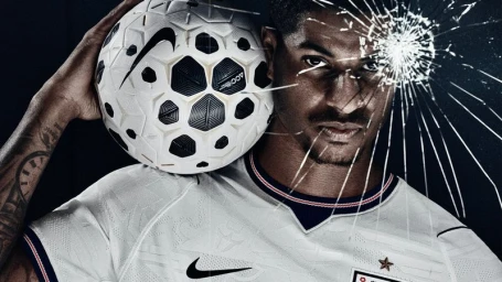 Marcus Rashford holding a Nike soccer ball on his shoulder behind cracked glass in an England national kit against a black background.
