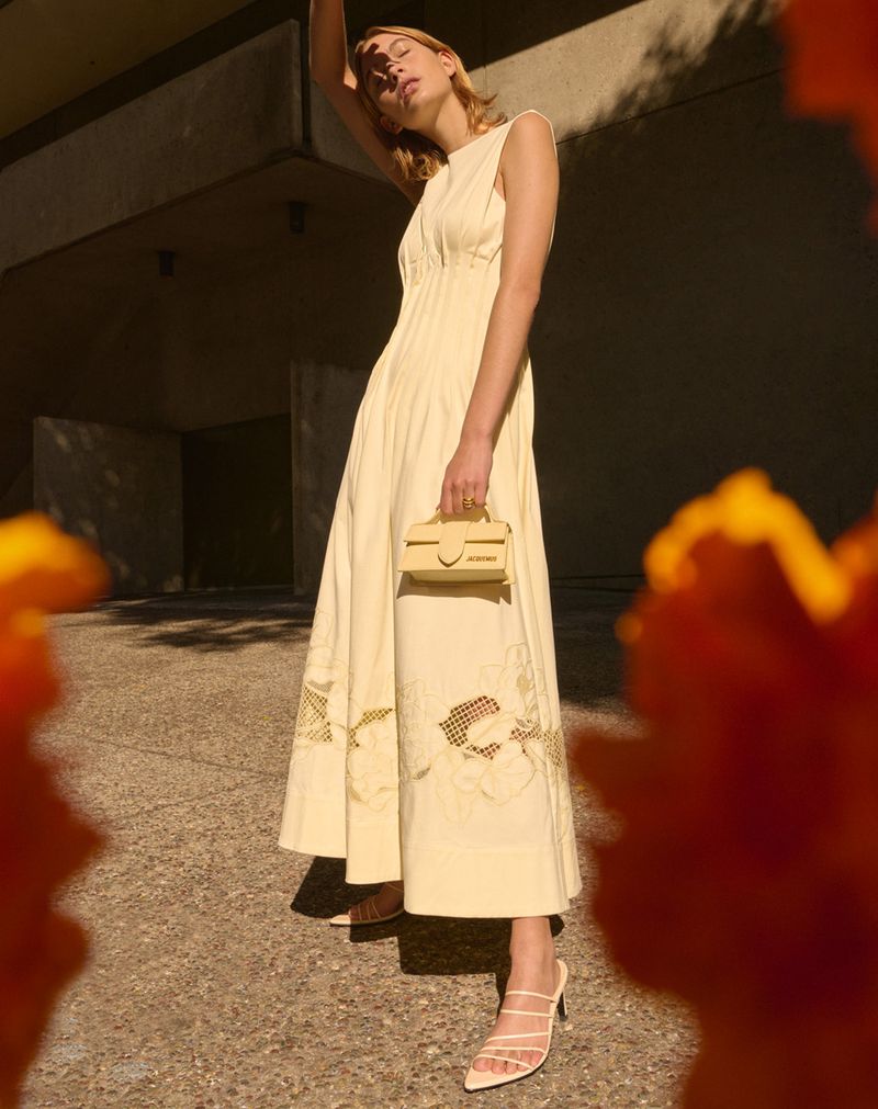 Adult woman in a sleeveless, floor-length yellow dress poses with a small yellow handbag in an urban outdoor setting with orange flowers.