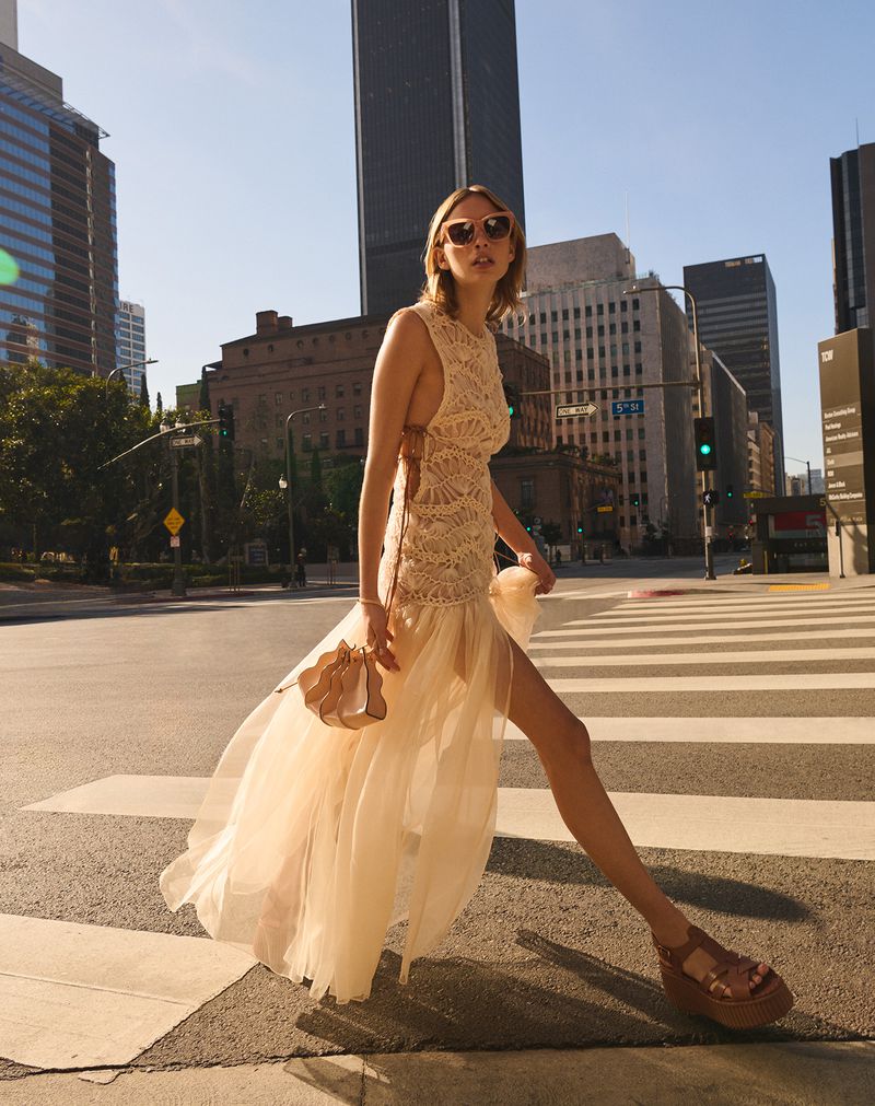 A woman in a light-colored gown crosses a city street on a sunny day, accessorized with sunglasses and a small handbag.