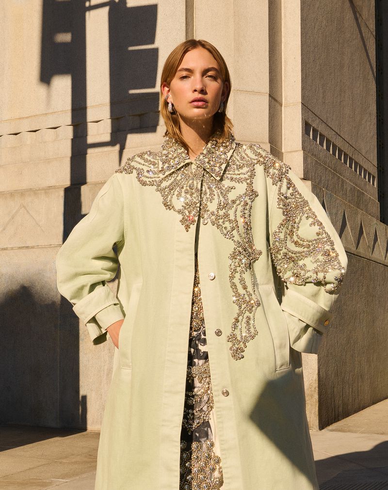 A woman poses outdoors wearing a long, light-green, heavily-ornamented coat and a patterned dress; sunlight and shadows highlight the architectural background.