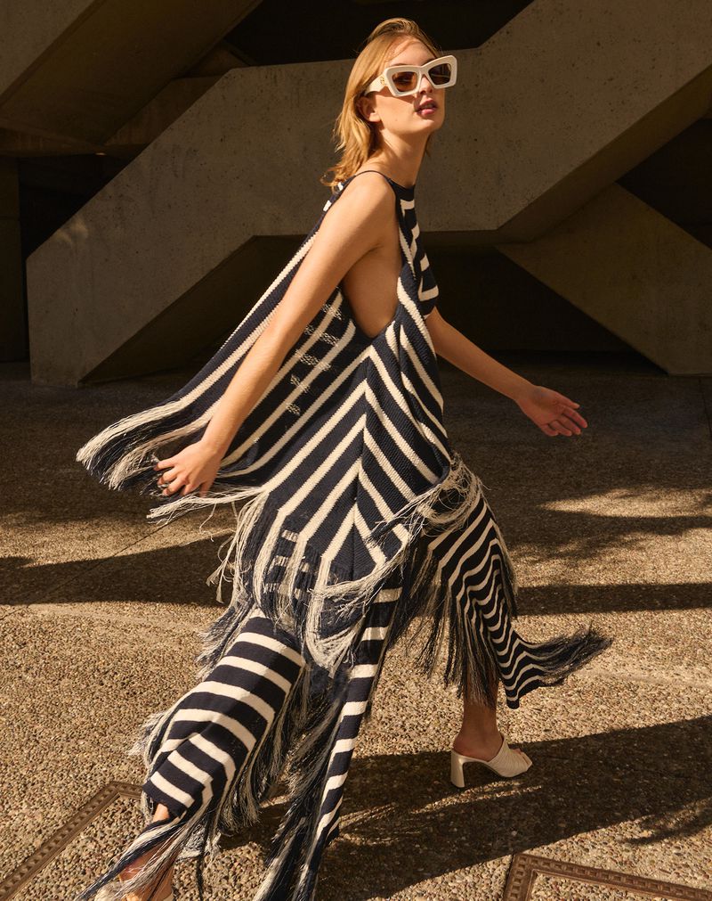 A blonde adult female model walks in a stylish black-and-white striped fringe dress and white sunglasses in an outdoor urban environment.