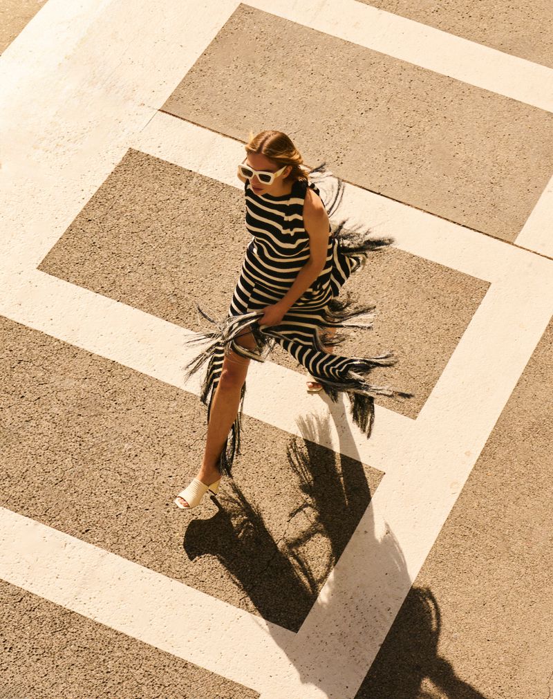 Eye-level shot of an adult woman in a black and white striped dress and white sunglasses posing outdoors on a patterned surface.