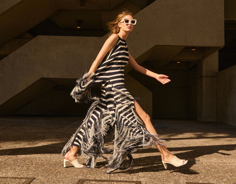 Fashionable woman walks outdoors in a striped halter dress, fringe detail, and square sunglasses, showcasing modern style and sophistication.