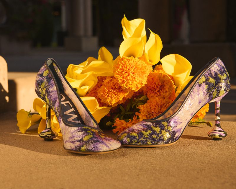 Pair of purple patterned high-heel shoes with yellow and orange flowers, illuminated by natural light on a neutral surface.