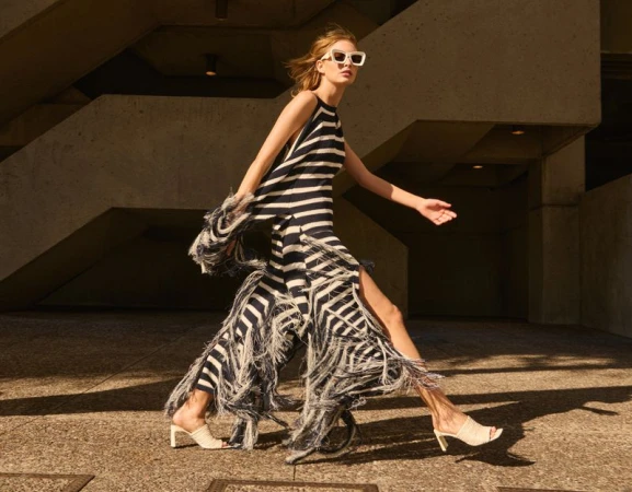 Model wearing a black and white striped fringe dress and white sunglasses walking outdoors in bright sunlight.