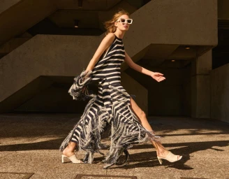 Model wearing a black and white striped fringe dress and white sunglasses walking outdoors in bright sunlight.