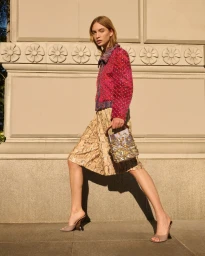 Model in magenta embellished jacket and patterned skirt holding a sequined handbag against an ornate stone building exterior.