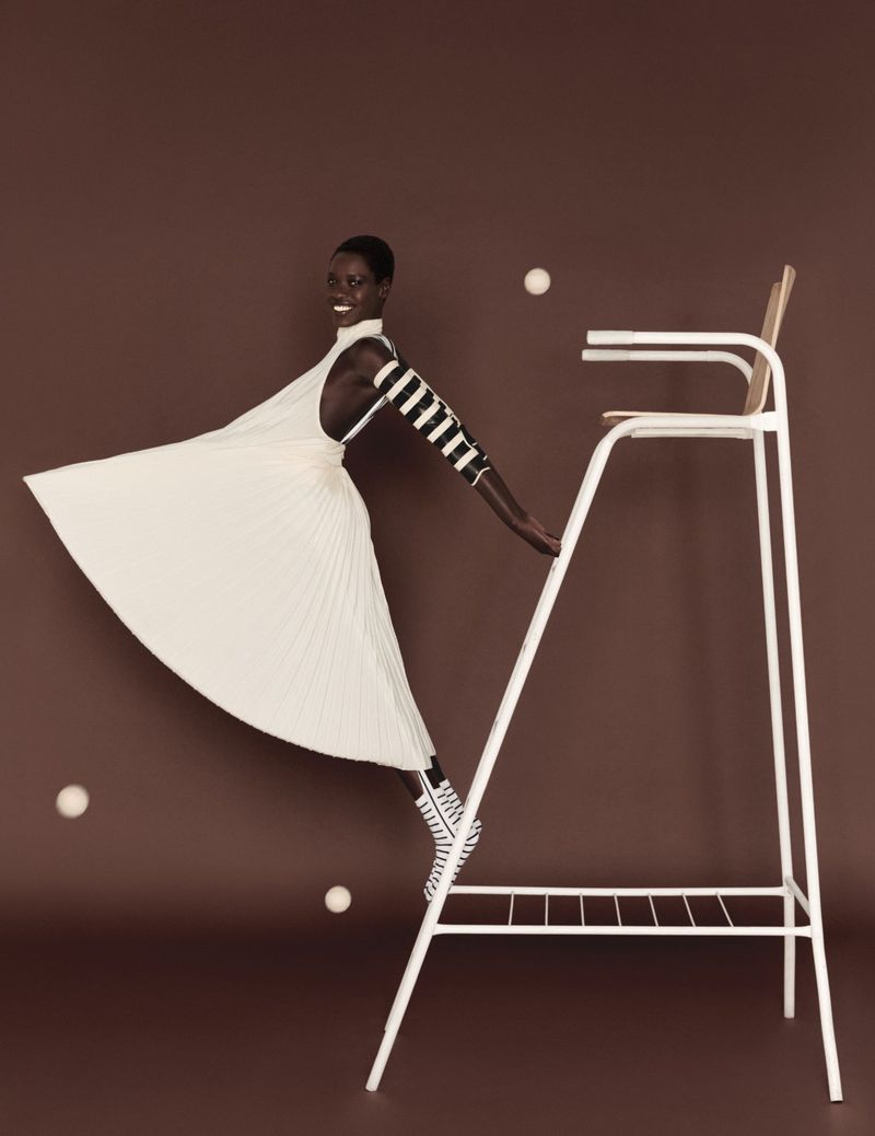 Model Awar Odhiang in a white pleated dress leaning off a tall umpire chair against a solid brown studio background.