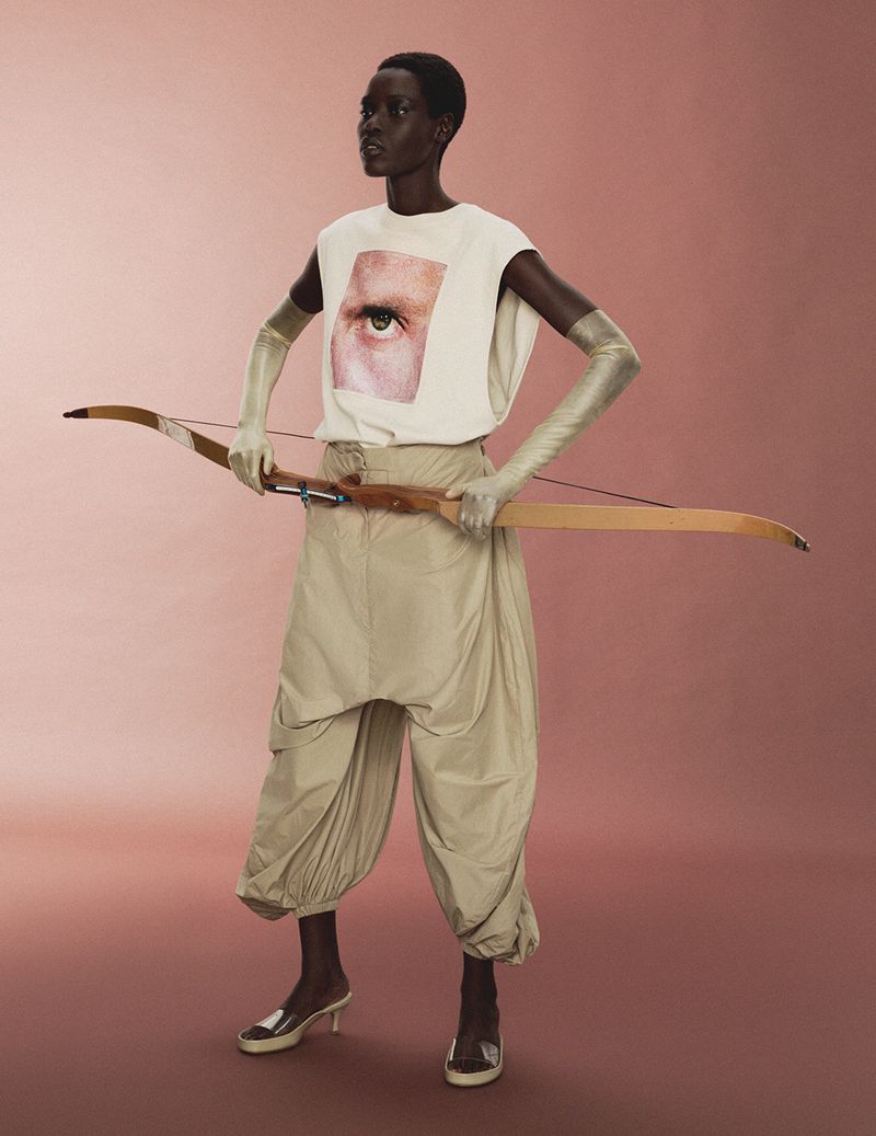 Model Awar Odhiang holding a wooden archery bow, wearing a graphic sleeveless top, draped khaki pants, and long gloves against a pink background.
