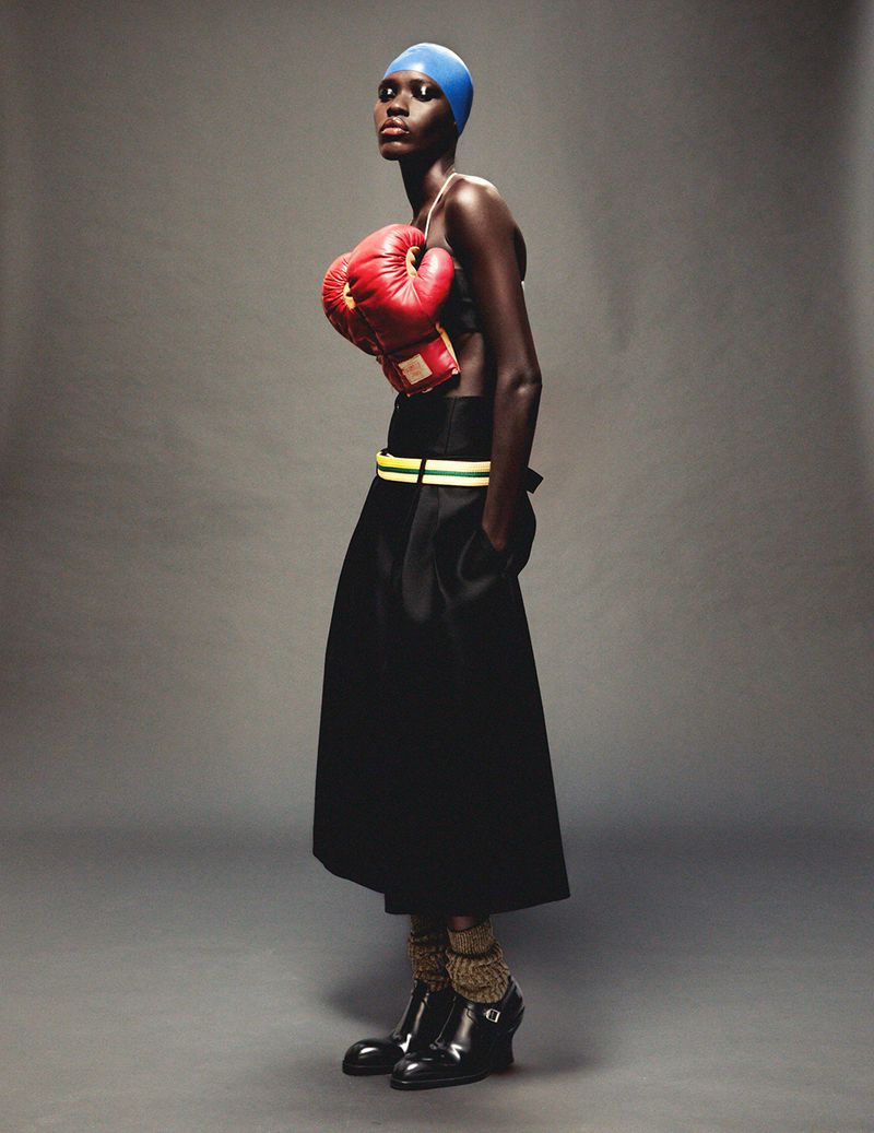 Model Awar Odhiang poses wearing a blue head cap, red boxing gloves strapped as a top, and a black skirt against a grey background.