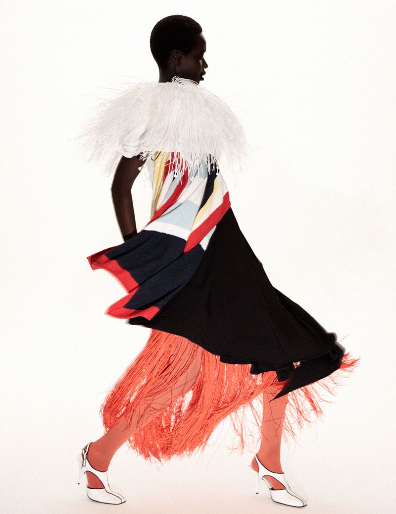 Model Awar Odhiang walking in a dynamic colorblock dress with a white feathered collar and orange fringe against a white backdrop.