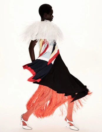Model Awar Odhiang walking in a dynamic colorblock dress with a white feathered collar and orange fringe against a white backdrop.