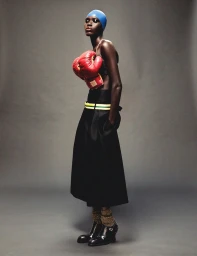 Model Awar Odhiang poses wearing a blue head cap, red boxing gloves strapped as a top, and a black skirt against a grey background.