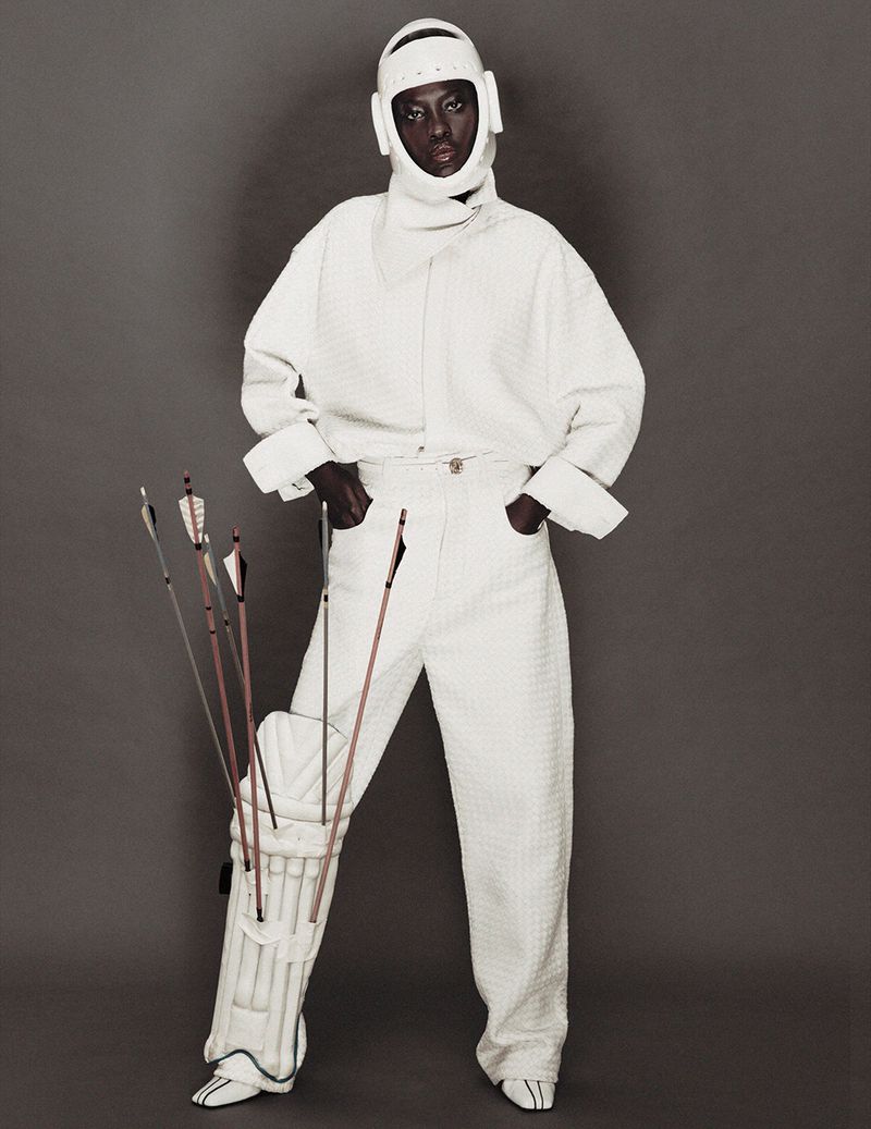 Model Awar Odhiang posing in an all-white textured outfit with a protective helmet and arrows tucked into a leg pad.