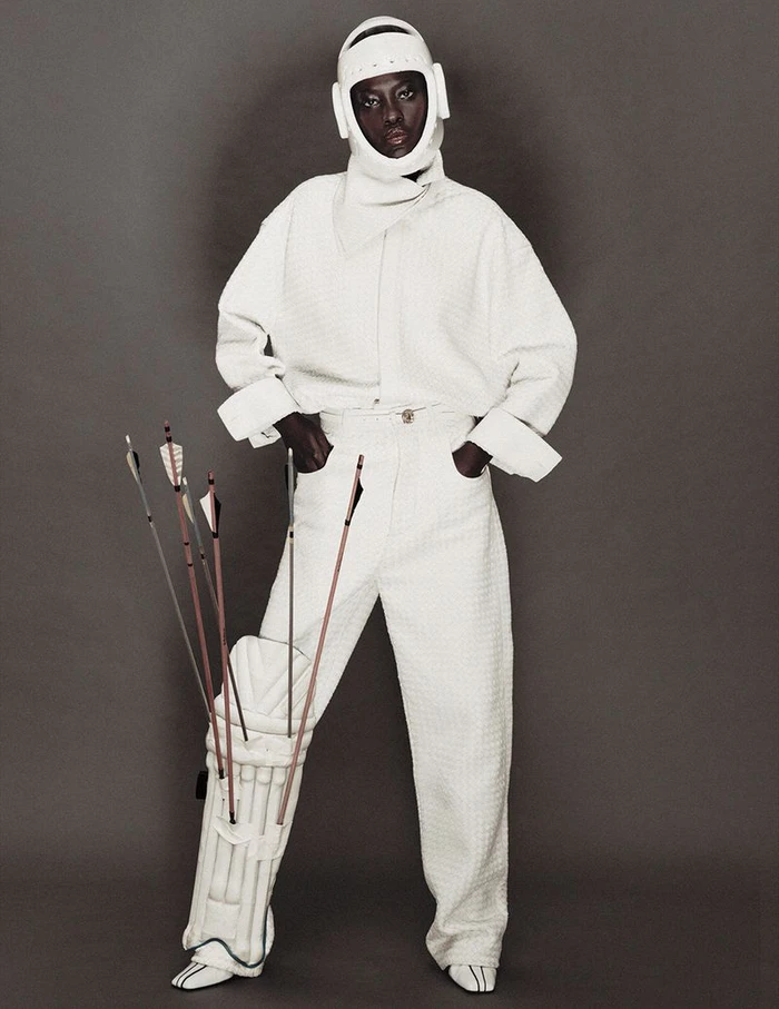 Model Awar Odhiang posing in an all-white textured outfit with a protective helmet and arrows tucked into a leg pad.