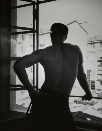Black and white photo of a shirtless man with his back turned, wearing a dark flat cap and looking out an open window.