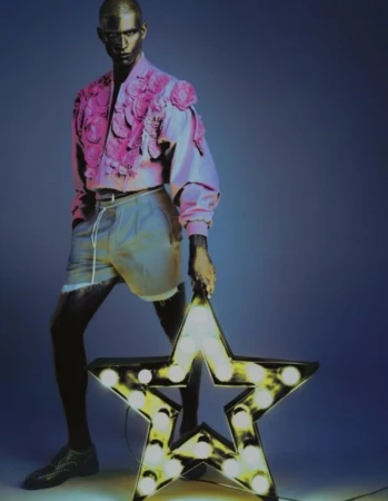Model Melvyn Reiningi in a pink floral appliqued jacket and shorts holding an illuminated marquee star against a blue backdrop.