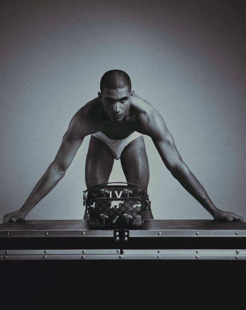 Model Melvyn Reiningi wearing white briefs leaning over a trunk with a patterned bag against a studio backdrop.
