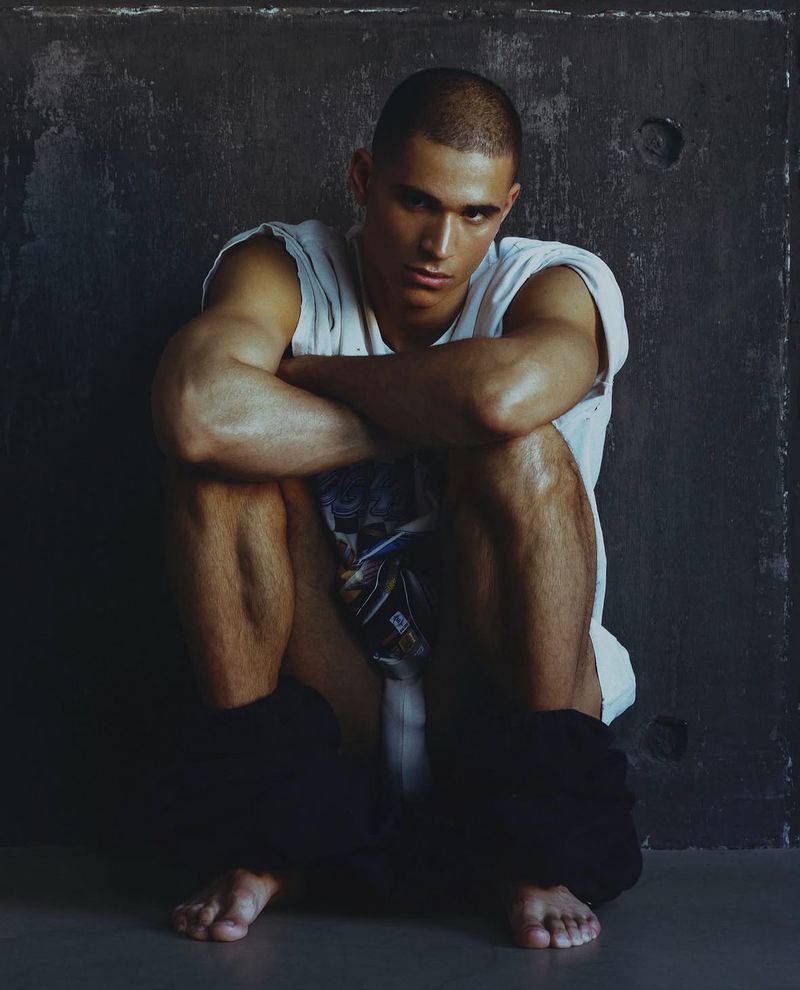 Model Melvyn Reiningi crouching barefoot in a white sleeveless t-shirt against a dark concrete wall for Numéro Homme Switzerland.
