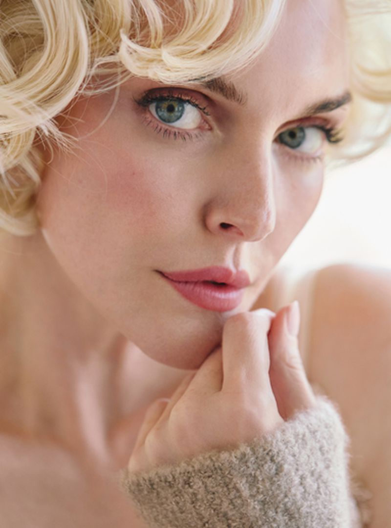 Close-up beauty portrait of Sophie Dahl with blonde curls and pink lipstick touching her chin in a knit sleeve.