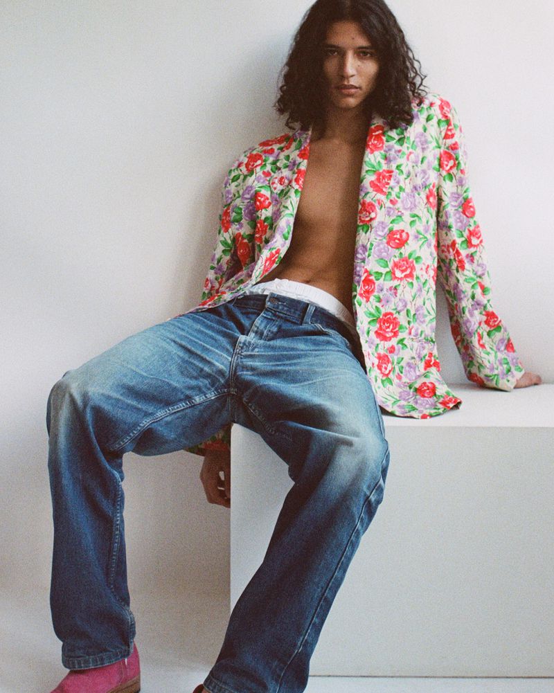 Model Skander Gueye wearing an open floral blazer and relaxed blue denim jeans seated on a white block against a white backdrop.