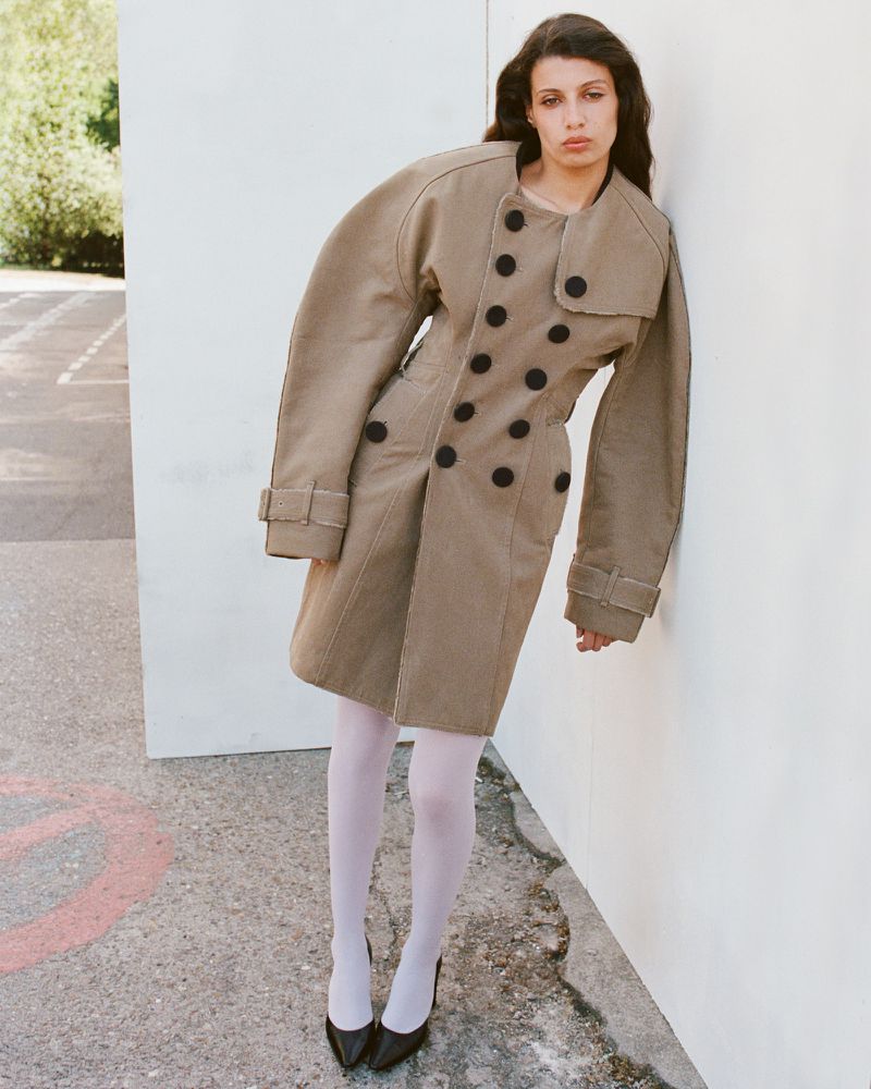 Model wearing a sculptural taupe trench coat and white tights leaning against a white outdoor panel set.