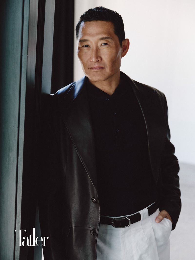 Daniel Dae Kim wearing a black leather blazer, black polo shirt, and white trousers leaning against a dark doorframe.