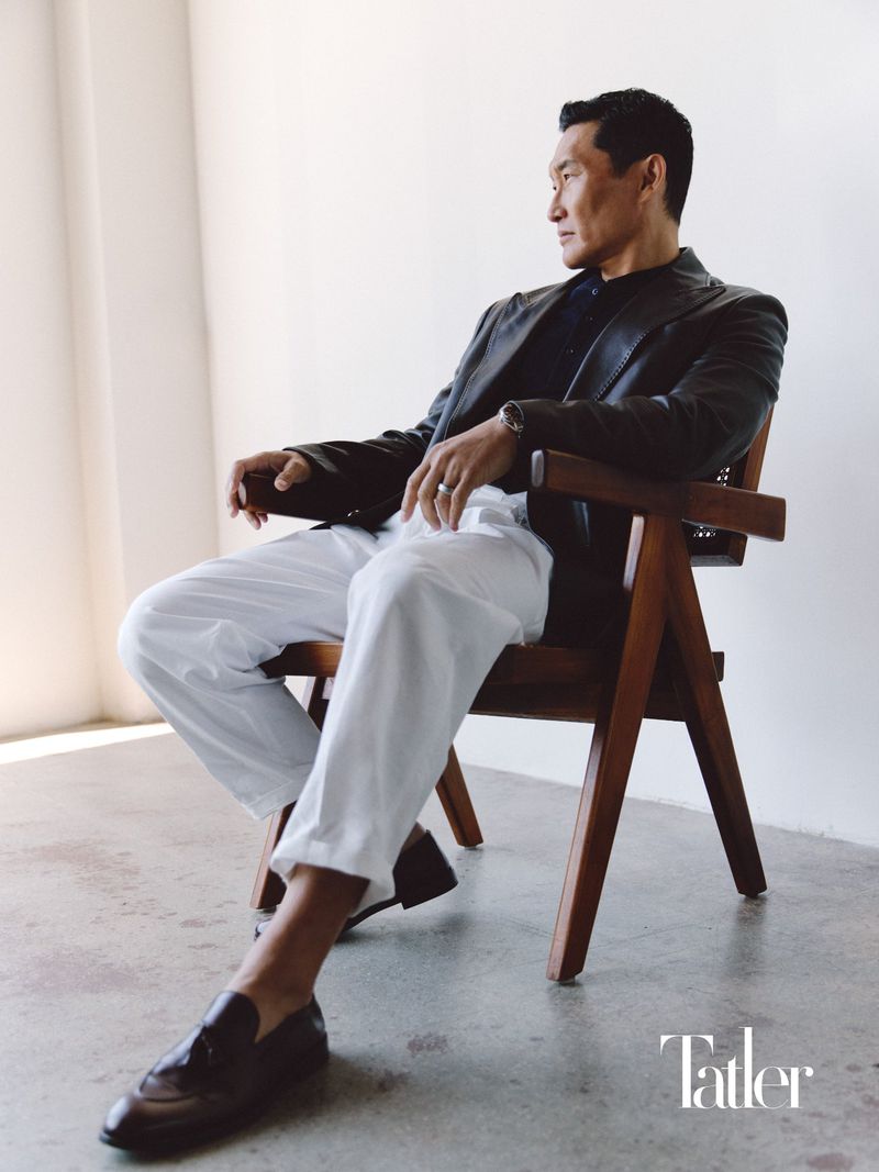 Actor Daniel Dae Kim seated in a wooden armchair wearing a dark leather jacket and white trousers for Tatler Asia.