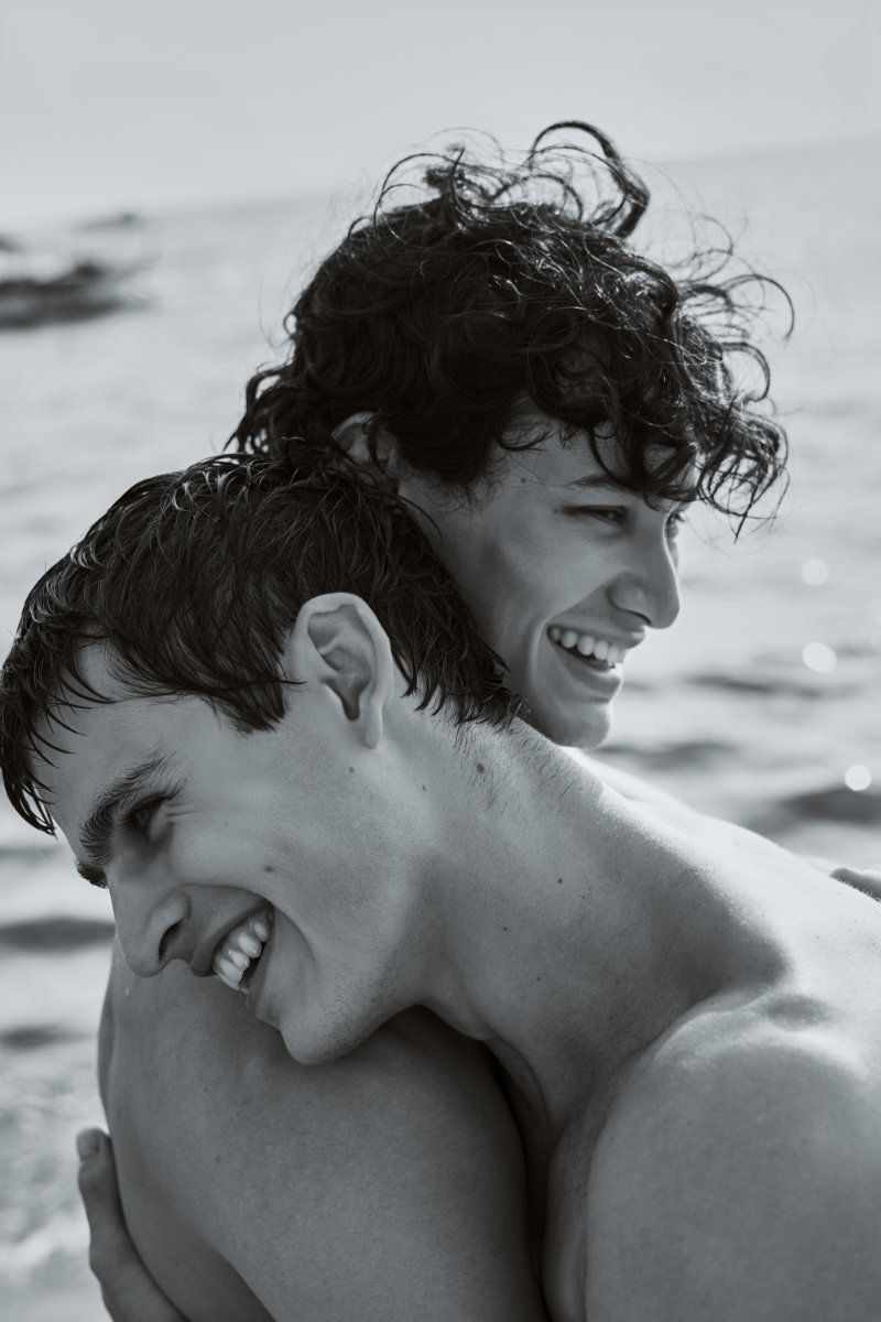 Close-up black and white portrait of two shirtless young men with wet hair smiling and embracing by the sea.