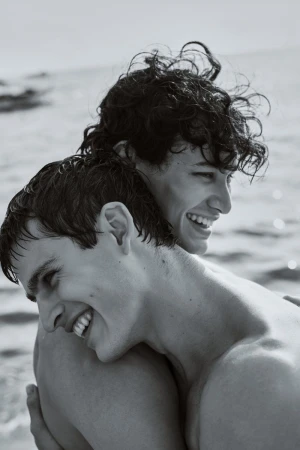 Close-up black and white portrait of two shirtless young men with wet hair smiling and embracing by the sea.