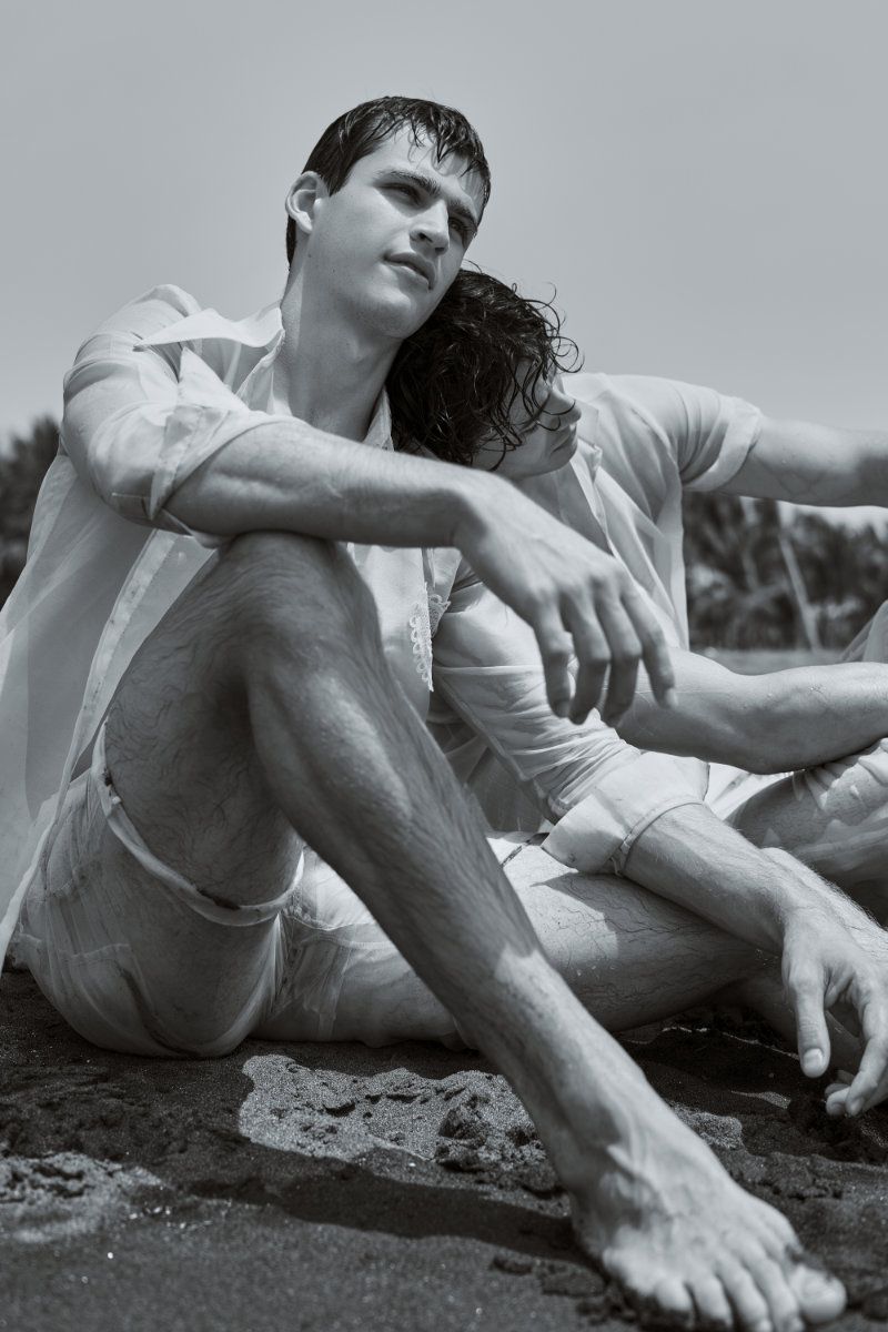 Two men in white shirts sitting close together on dark sand at the beach in a black and white editorial portrait.