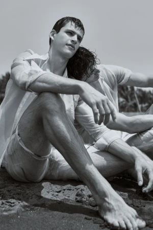 Two men in white shirts sitting close together on dark sand at the beach in a black and white editorial portrait.