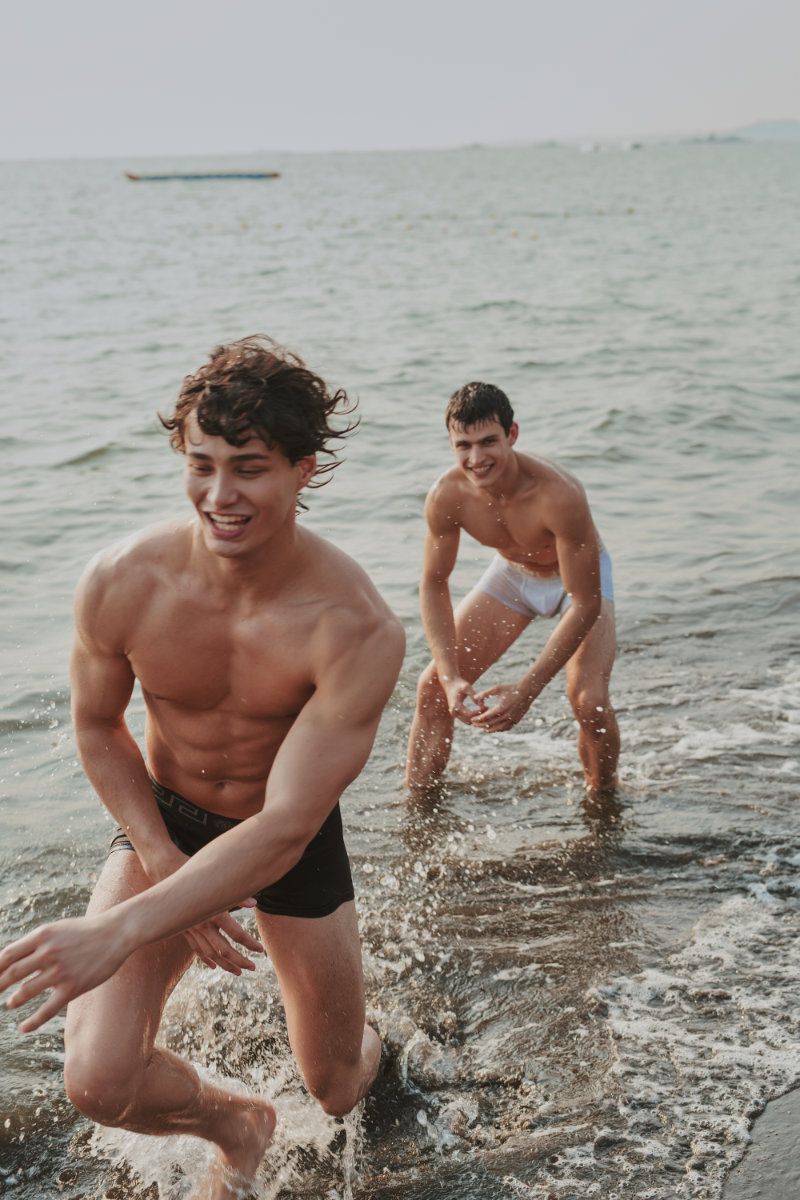 Two shirtless men in swim trunks running and splashing through ocean water towards the shore.