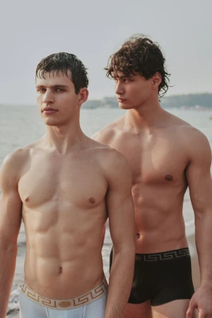 Two shirtless male models posing outdoors on a sandy beach wearing patterned waistband boxer briefs.