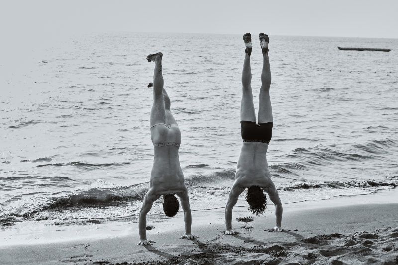 Black and white photo of two men performing handstands on a wet sandy beach with ocean waves in the background.