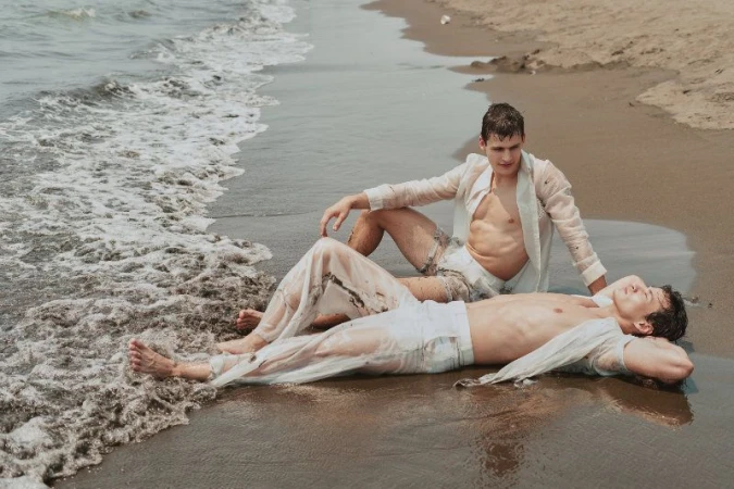 Two male models in wet white linen shirts and trousers lying on a wet sandy beach near the ocean waves