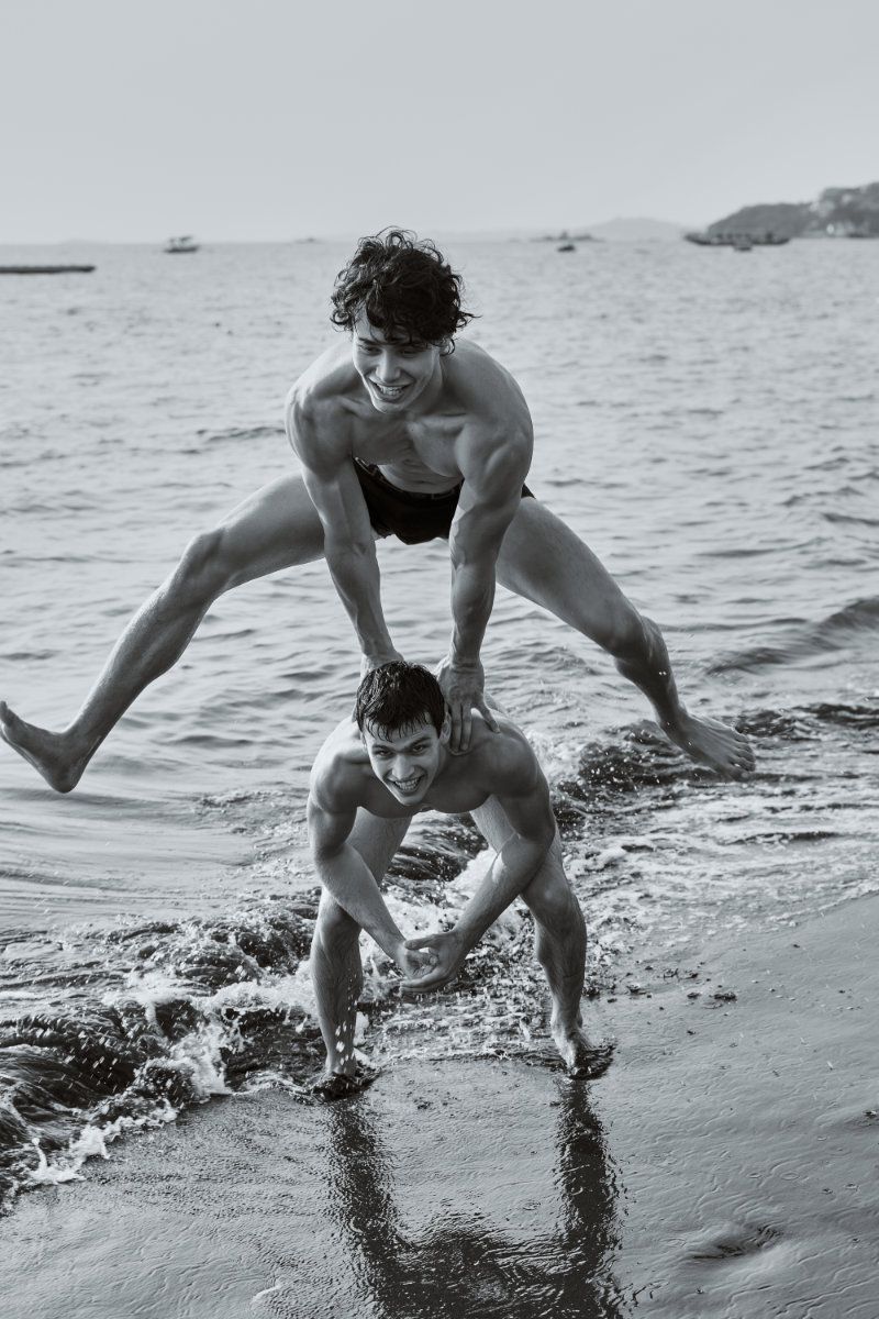 Black and white photo of a shirtless man leaping mid-air over another crouching man on a wet sandy beach shoreline.