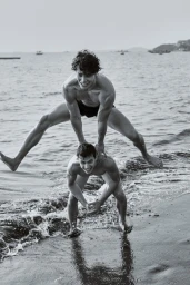 Black and white photo of a shirtless man leaping mid-air over another crouching man on a wet sandy beach shoreline.