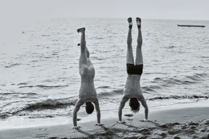 Black and white photo of two men performing handstands on a wet sandy beach with ocean waves in the background.