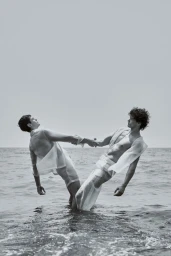 Two men in sheer white clothing hold hands while leaning away from each other in ocean water.