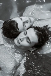 Black and white overhead portrait of two young men floating close together in rippling water with their eyes closed.