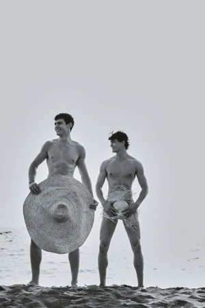 Two shirtless men standing on a sandy beach holding large woven straw hats in a black and white editorial.