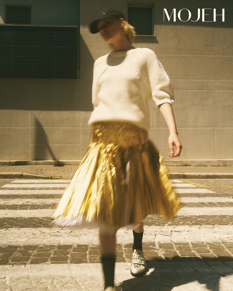 Woman walking across a cobblestone crosswalk wearing a white short-sleeve knit top, metallic gold pleated skirt, and a black cap.