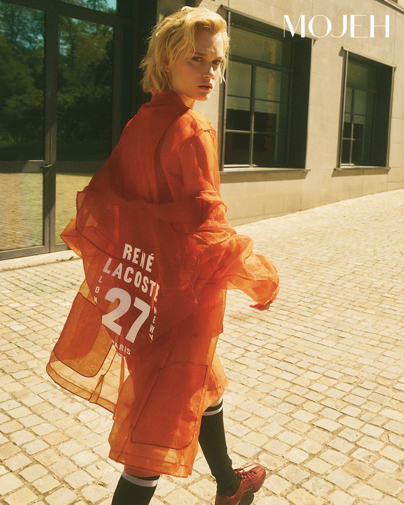 Model wearing a sheer orange coat with white graphic lettering looking over her shoulder on a paved stone courtyard.
