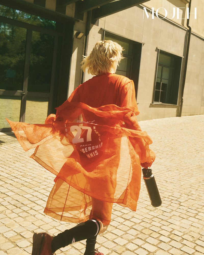 Blonde woman seen from behind running in a sheer orange jacket and black socks across a sunny cobblestone courtyard.